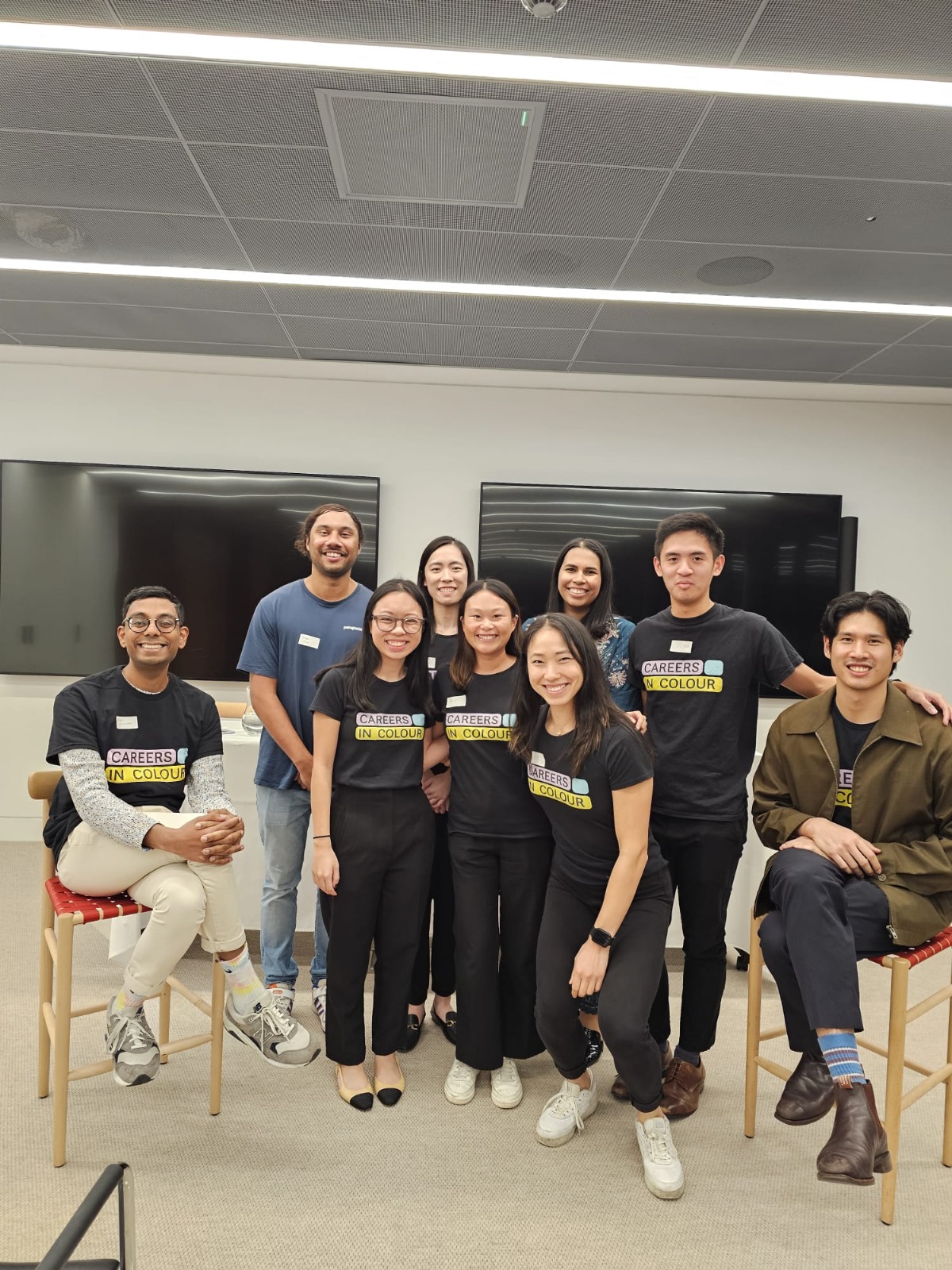 Group of young professionals posing together indoors, many wearing matching “Careers in Colour” t-shirts, smiling at the camera in a modern office space.